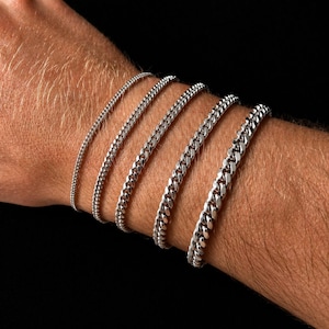 Puede incluir: Una colección de pulseras de cadena plateadas de diferentes anchos se muestra en una muñeca. Las pulseras presentan diferentes estilos de eslabones, que van desde delicados hasta gruesos. El fondo es negro liso, resaltando las joyas.