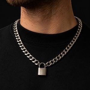 May include: A silver-toned curb chain necklace featuring a polished metal padlock pendant. The thick, interlocking links of the chain connect to the base of the padlock, which hangs centered at the bottom of the necklace against a black shirt.