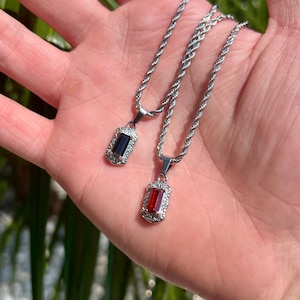 Könnte beinhalten: Zwei Halsketten aus Edelstahl mit rechteckigen Anhängern. Ein Anhänger hat einen dunkelblauen Stein, der andere einen roten Stein, beide umgeben von einem dekorativen Rand. Die Ketten haben ein seilartiges Design.