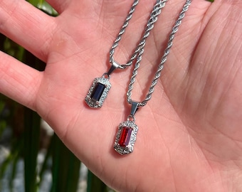 May include: Two silver-coloured necklaces, each with a rectangular pendant. One pendant has a dark blue stone, the other a red stone, both with a decorative border. The chains have a twisted rope design.