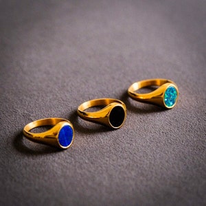 Peut inclure: Trois bagues signet en or avec des pierres de couleurs différentes. Les bagues ont un dessus rond et plat avec une finition lisse et polie. Les pierres sont noires, bleues et bleu clair.