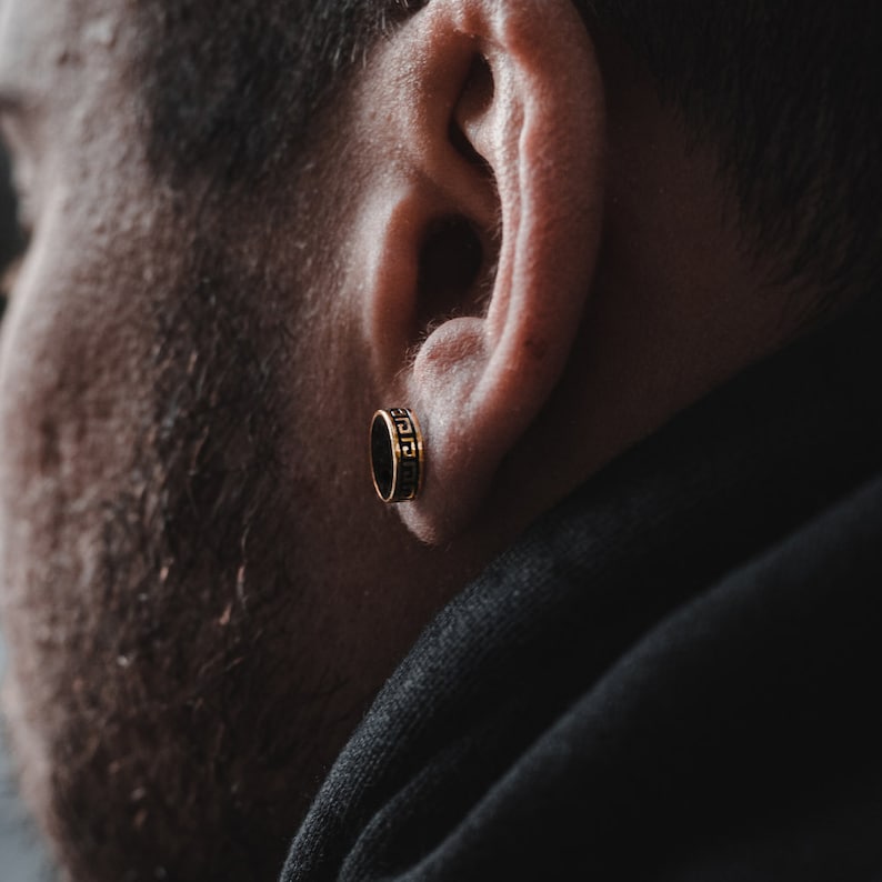 May include: A close-up of a person's ear with a gold earring featuring a black and gold geometric design.