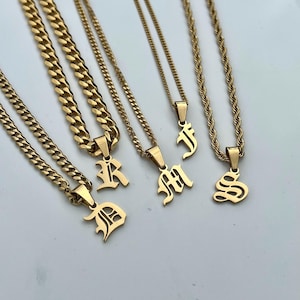May include: A selection of gold-coloured initial pendant necklaces. The necklaces feature different chain styles, including curb and rope chains. Each pendant displays a letter in a gothic font. The letters visible are D, R, M, J, and S.