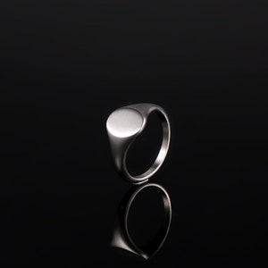 May include: A silver signet ring with a smooth, oval-shaped top. The ring is on a black background.