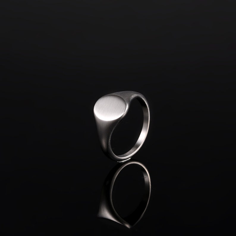 May include: A silver signet ring with a smooth, oval-shaped top. The ring is on a black background.