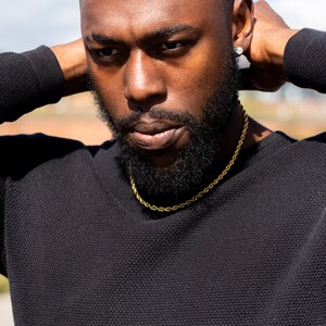 Puede incluir: Primer plano de una persona que lleva un su&eacute;ter negro con un collar de cadena de cuerda dorada. La persona tiene barba y mira hacia abajo.