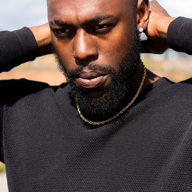 Puede incluir: Primer plano de una persona que lleva un su&eacute;ter negro con un collar de cadena de cuerda dorada. La persona tiene barba y mira hacia abajo.