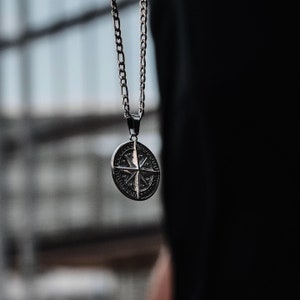 K&ouml;nnte beinhalten: Eine silberne Kettenkette mit einem Kompass-Anh&auml;nger. Der Kompass hat eine silberne Oberfl&auml;che und zeigt ein detailliertes Design einer Kompassrose mit Himmelsrichtungen.