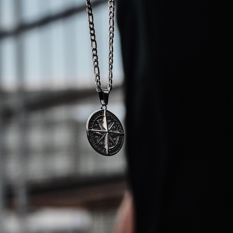 K&ouml;nnte beinhalten: Eine silberne Kettenkette mit einem Kompass-Anh&auml;nger. Der Kompass hat eine silberne Oberfl&auml;che und zeigt ein detailliertes Design einer Kompassrose mit Himmelsrichtungen.