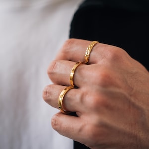 May include: Three gold-toned rings worn on three fingers. The top ring features a textured, hammered surface. The middle ring has a smooth, flat band. The bottom ring displays a polished, rounded surface with subtle vertical detailing along the edges.