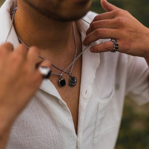 May include: Silver necklaces with pendant charms, including a black square and a circular design. The image also shows a silver ring with black accents. The jewelry is displayed against a white shirt.
