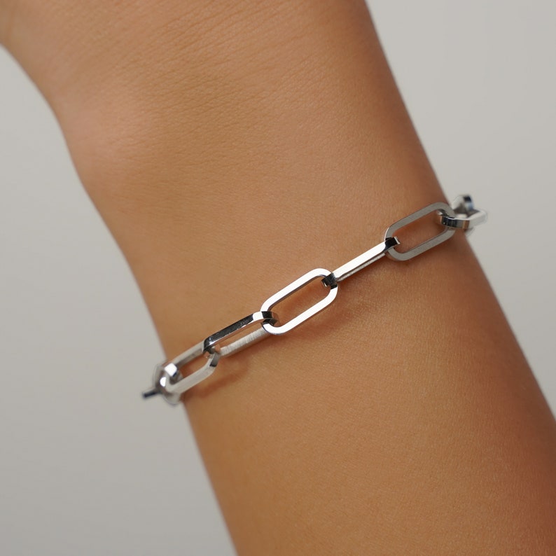 May include: A silver-colored link bracelet. The bracelet features a series of elongated, rectangular links connected to each other. The bracelet is worn on a wrist, and the links are visible against the skin.