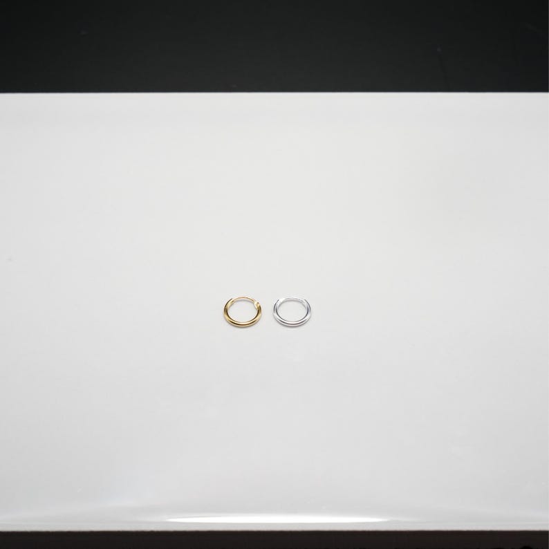 Puede incluir: Dos peque&ntilde;os pendientes de aro. Uno es de color dorado y el otro plateado. Ambos pendientes son circulares y parecen estar hechos de metal. Los pendientes se muestran sobre una superficie blanca.
