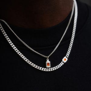 May include: Two silver necklaces with a cuban link chain design. One necklace features a small rectangular orange pendant, and the other has an orange rectangular gem on the chain. The necklaces are worn against a black top.