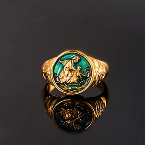 May include: A gold-toned ring featuring a circular face with a raised, metallic gold relief of a figure interacting with a smaller figure, set against a vibrant green enamel background. The band displays intricate, textured, wing-like side detailing.