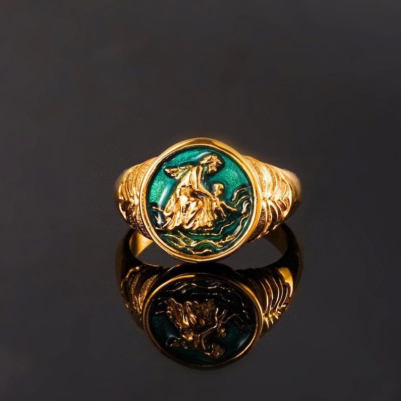 May include: A gold-toned ring featuring a circular face with a raised, metallic gold relief of a figure interacting with a smaller figure, set against a vibrant green enamel background. The band displays intricate, textured, wing-like side detailing.