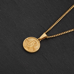 Könnte beinhalten: Eine goldfarbene Metallhalskette mit einem runden Anhänger, der ein erhabenes Relief einer Figur mit einem Stab zeigt. Der Anhänger ist an einer feinen Panzerkette befestigt und besticht durch eine polierte Oberfläche und feine Details.
