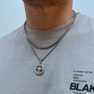 May include: A silver-toned necklace set featuring a chain and a pendant. The pendant is a compass design with a star in the center and surrounding clear stones. The person is wearing a gray t-shirt with the word "BLAK" printed on it.