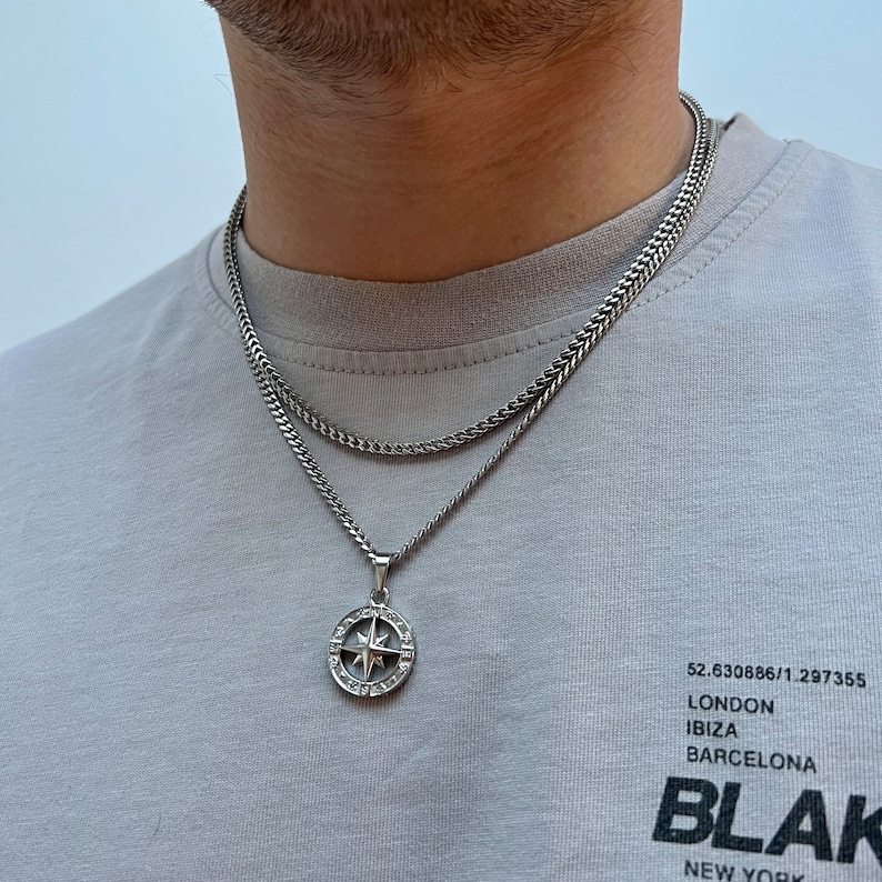 May include: A silver-toned necklace set featuring a chain and a pendant. The pendant is a compass design with a star in the center and surrounding clear stones. The person is wearing a gray t-shirt with the word "BLAK" printed on it.