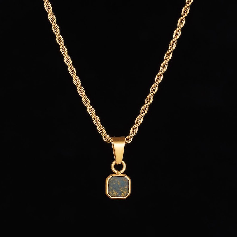 May include: Gold-toned necklace with a square pendant. The pendant has a dark blue-green center with gold flecks, set in a gold frame. The chain is a twisted rope design, and the background is black.