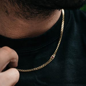 May include: A gold-toned curb chain necklace featuring a single rectangular link set with a small, deep red gemstone. The chain has a classic interlocking link design and rests against a plain black fabric background, highlighting the metallic finish.