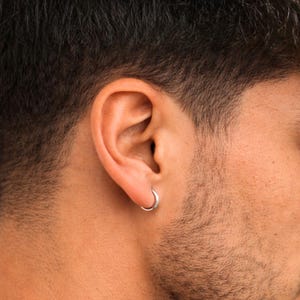 Op de afbeelding: Close-up van een zilveren oorring die in een oor piercing wordt gedragen. De oorring is een kleine, eenvoudige cirkel. De huid van de persoon is zichtbaar, met donker haar en een baard.