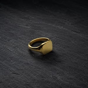 May include: A gold-toned signet ring featuring a smooth, flat, square-shaped face and a polished band. The ring has a reflective metallic finish and a solid construction, showcasing a clean, unadorned design with a uniform golden color throughout.