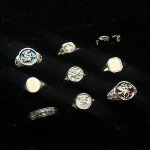 Könnte beinhalten: Eine Sammlung silberfarbener Ringe mit verschiedenen Designs, darunter ein runder Ring mit blauer Emaille, ein Kreuz-Siegelring, ein schlichter ovaler Siegelring, ein texturiertes Band mit griechischem Muster und ein roter Emaille-Ring mit Engelsmotiv.