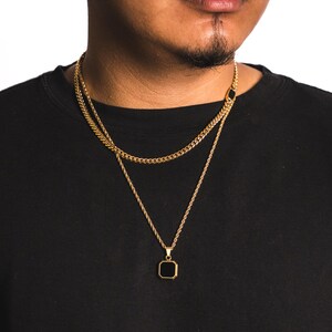 May include: Gold-toned layered necklace set. The top chain is a curb link style, and the bottom chain has a pendant with a black square stone. The chains are displayed against a black background.