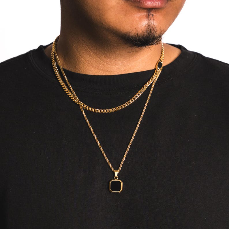 May include: Gold-toned layered necklace set. The top chain is a curb link style, and the bottom chain has a pendant with a black square stone. The chains are displayed against a black background.
