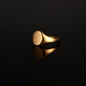 May include: A gold signet ring featuring a smooth, circular flat top surface and a polished band. The ring has a warm metallic finish and is captured with a clear reflection on the surface below, highlighting its simple, rounded design.