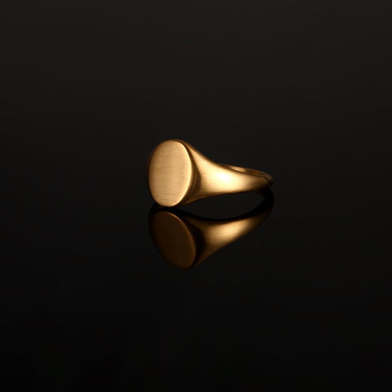 May include: A gold signet ring featuring a smooth, circular flat top surface and a polished band. The ring has a warm metallic finish and is captured with a clear reflection on the surface below, highlighting its simple, rounded design.