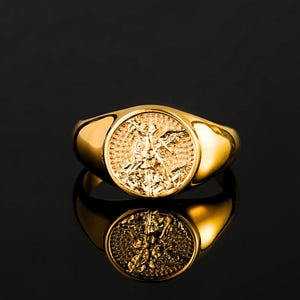 May include: A gold-toned signet ring featuring a circular face with an embossed relief of a winged figure holding a sword. The ring has a polished, smooth band and a reflective surface, showcasing intricate detailing on the central coin-like design.