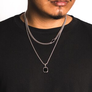 May include: Two silver necklaces. The top necklace is a curb chain, and the bottom necklace has a black square pendant. The necklaces are worn on a person wearing a black shirt.