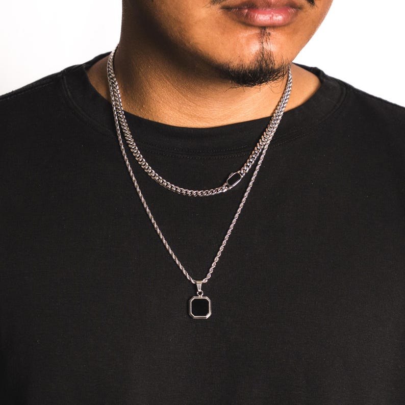 May include: Two silver necklaces. The top necklace is a curb chain, and the bottom necklace has a black square pendant. The necklaces are worn on a person wearing a black shirt.