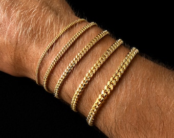 May include: Five gold chain bracelets of varying widths are displayed on a person's wrist against a black background. The bracelets have a curb link design, with a polished finish. The bracelets are different sizes, from delicate to bold.