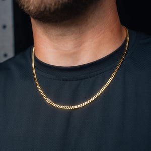 May include: A gold-toned curb chain necklace featuring a single rectangular link accented with small, clear, sparkling stones. The chain rests against a dark-colored shirt, showcasing a polished metallic finish and a classic interlocking link design.