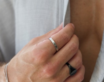 Peut inclure: Une main portant deux bagues. La bague sur l'index est une fine bande argentée. La bague sur l'auriculaire est une chevalière noire.
