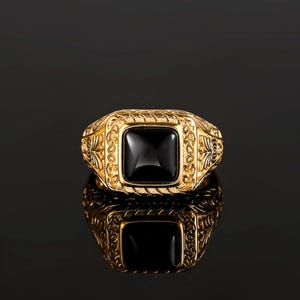May include: A gold ring featuring a square-cut black stone set in the center. The band and the setting are decorated with intricate, raised floral and leaf-like engravings that extend along the sides of the ring, creating a textured appearance.