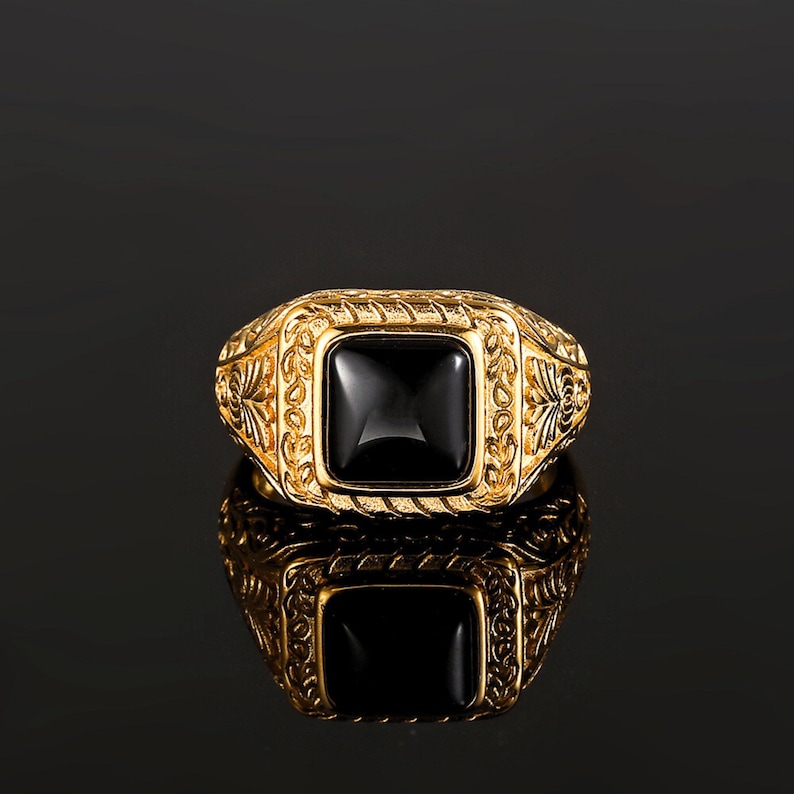 May include: A gold ring featuring a square-cut black stone set in the center. The band and the setting are decorated with intricate, raised floral and leaf-like engravings that extend along the sides of the ring, creating a textured appearance.