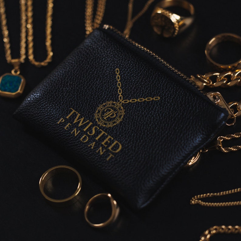 May include: A black leather pouch with a gold zipper and the words "Twisted Pendant" printed on the front. The pouch is surrounded by gold chains and rings.