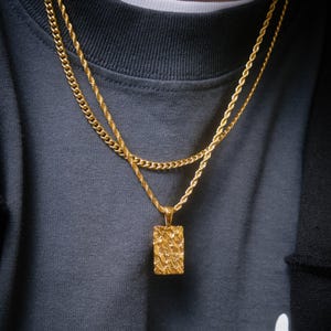 May include: Two gold chains with a textured rectangular pendant. The chains are layered on top of each other and the pendant is hanging from the bottom chain.