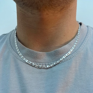 May include: A silver mariner chain necklace worn on a person's neck. The chain is made of small, square links.