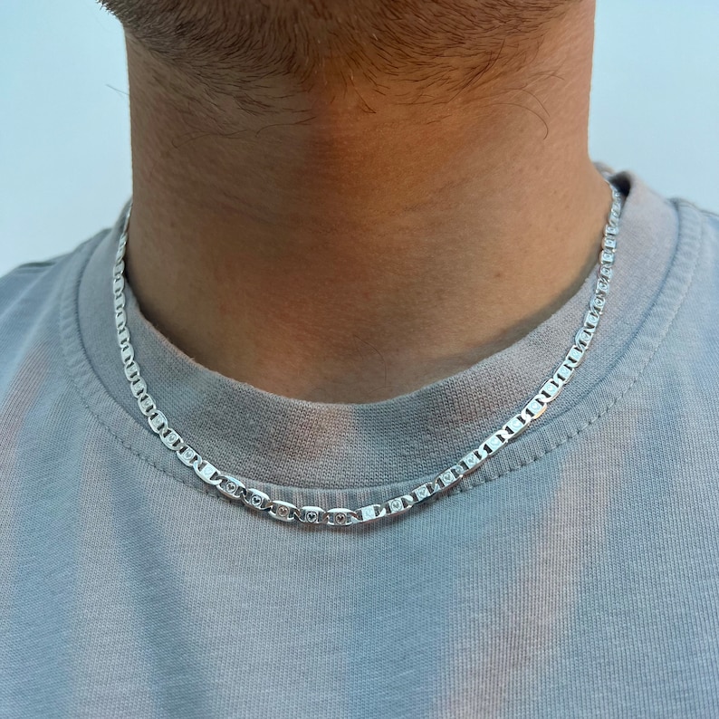 May include: A silver mariner chain necklace worn on a person's neck. The chain is made of small, square links.