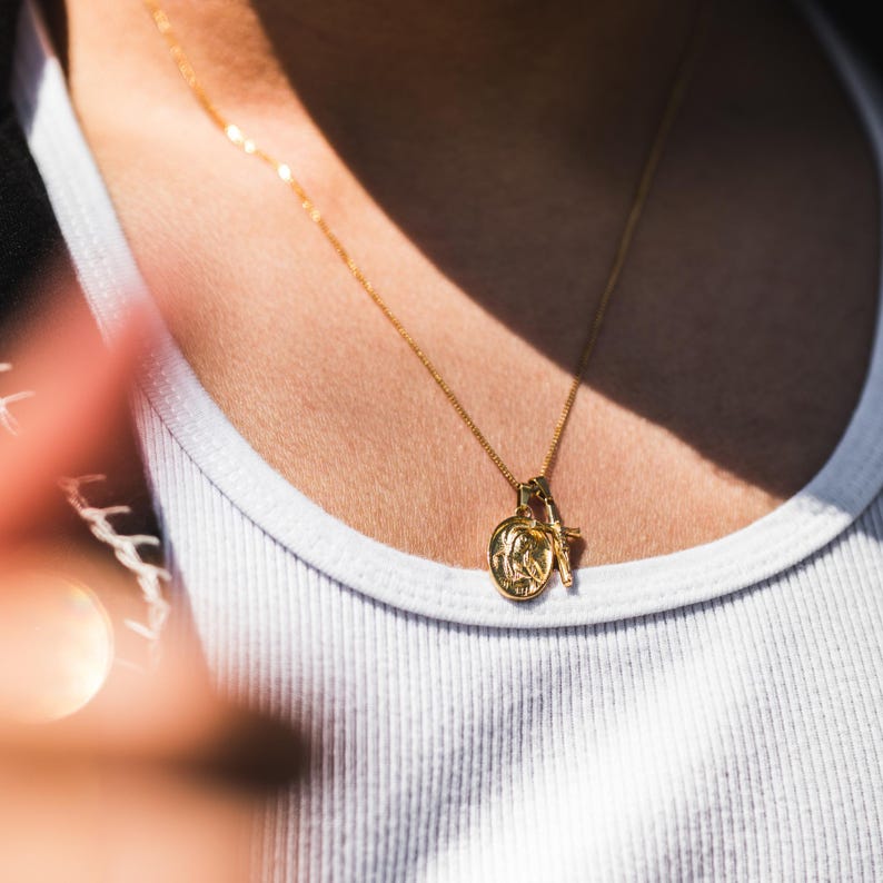 May include: A gold-colored necklace with a pendant featuring a circular design and a small cross. The necklace is worn over a white tank top. The image is well-lit, highlighting the jewelry's details.