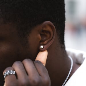 May include: A close-up of a person's ear with a small, round, silver earring. The person's finger is touching their earlobe.