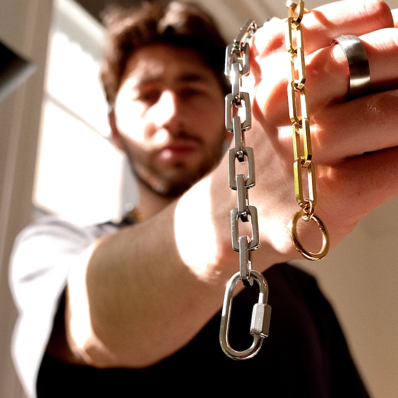 May include: Two chain necklaces, one silver and one gold, are held up in front of a person. The silver chain has a carabiner clasp. The gold chain has a circular clasp. A silver ring is on the person's finger.