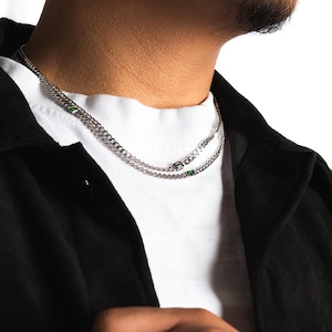 May include: A silver chain necklace with two strands, each featuring small, square green accents. The necklace is worn over a white t-shirt and a black jacket. The image is a close-up of the neck and upper chest.