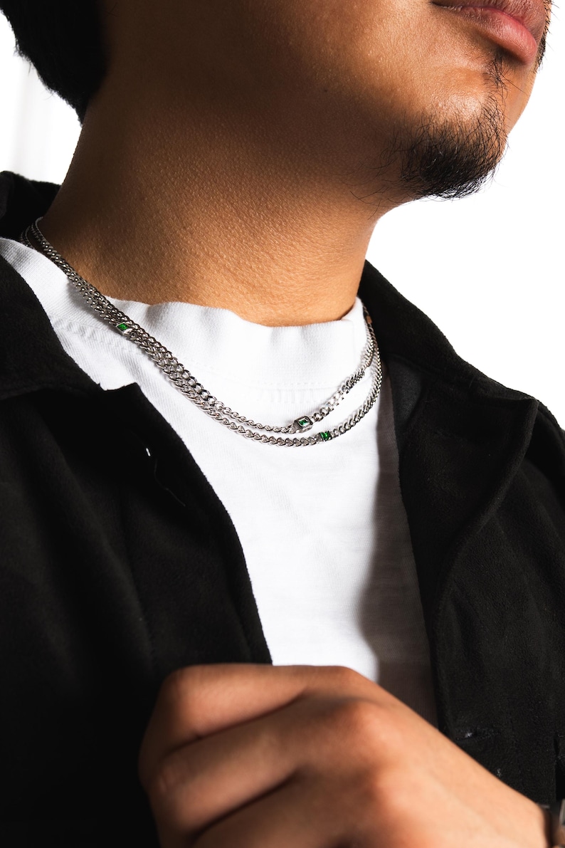 May include: A silver chain necklace with two strands, each featuring small, square green accents. The necklace is worn over a white t-shirt and a black jacket. The image is a close-up of the neck and upper chest.