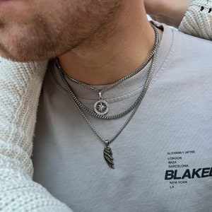 May include: Three silver necklaces with different pendants. The top necklace has a round pendant with a compass design. The middle necklace is a thicker chain. The bottom necklace has a wing pendant. The person is wearing a gray t-shirt.